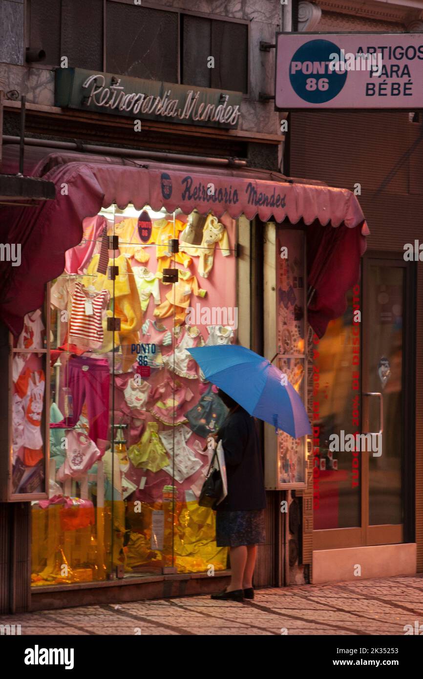 Une photo verticale d'une personne avec un parapluie bleu regardant un magasin de vêtements pour bébés à Sesimbra, Portugal Banque D'Images
