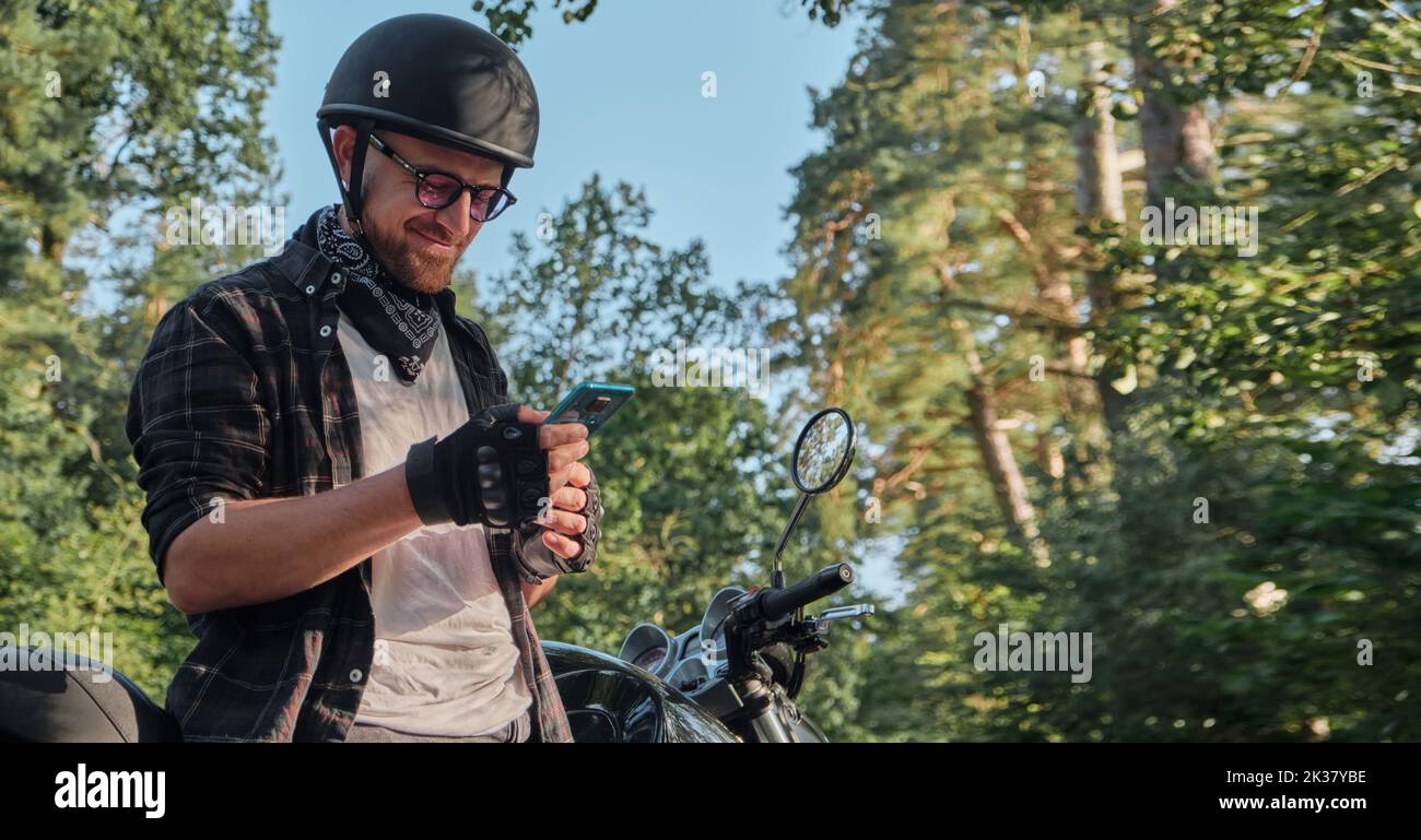Jeune homme motard dans un casque avec un téléphone portable et souriant assis sur une moto Banque D'Images