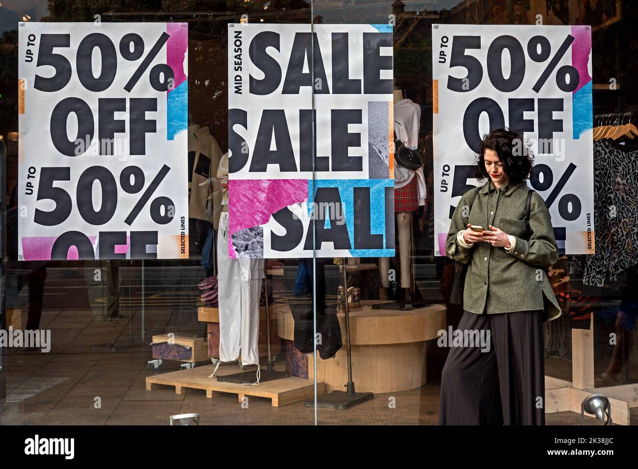 Jeune femme regardant son téléphone avec 50% de rabais sur les affiches de solde sur la fenêtre du magasin derrière elle. Édimbourg, Écosse, Royaume-Uni. Banque D'Images