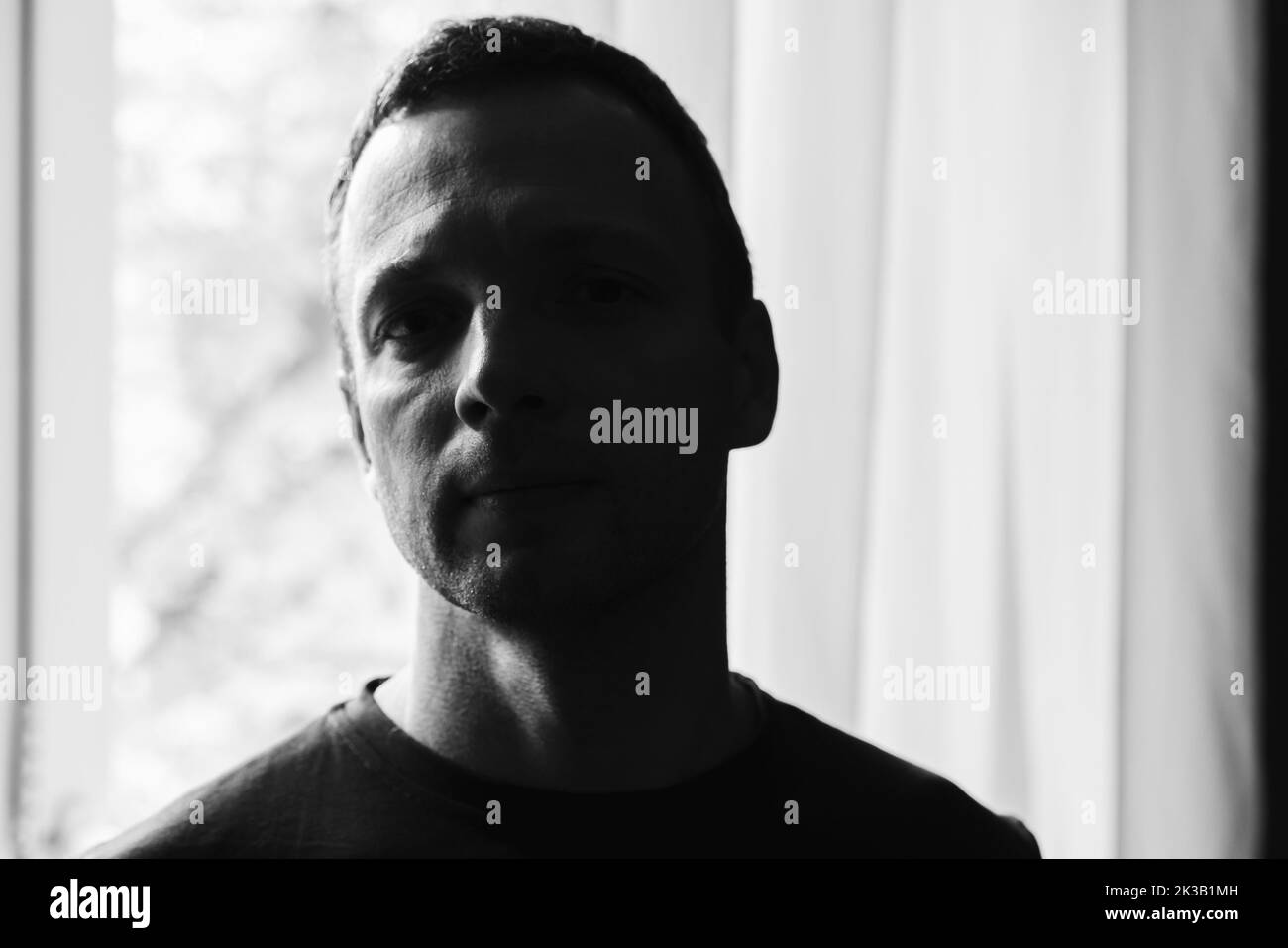 Portrait artistique en studio sombre d'un jeune homme caucasien adulte debout devant une fenêtre lumineuse, photo en noir et blanc avec une mise au point sélective douce Banque D'Images