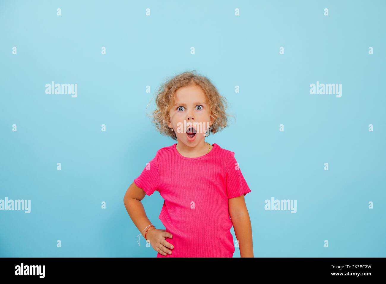 Portrait d'une petite fille choquée avec des cheveux bouclés et équitables portant un T-shirt rose, avec une bouche ouverte sur fond bleu. Banque D'Images