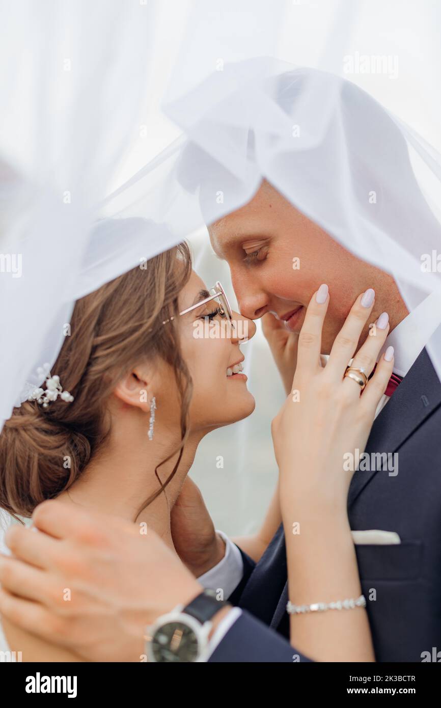 Vue latérale d'un couple souriant debout sous un voile de mariée. Jeune femme touchant le visage, jeune marié élégant qui embrasse la mariée. Banque D'Images