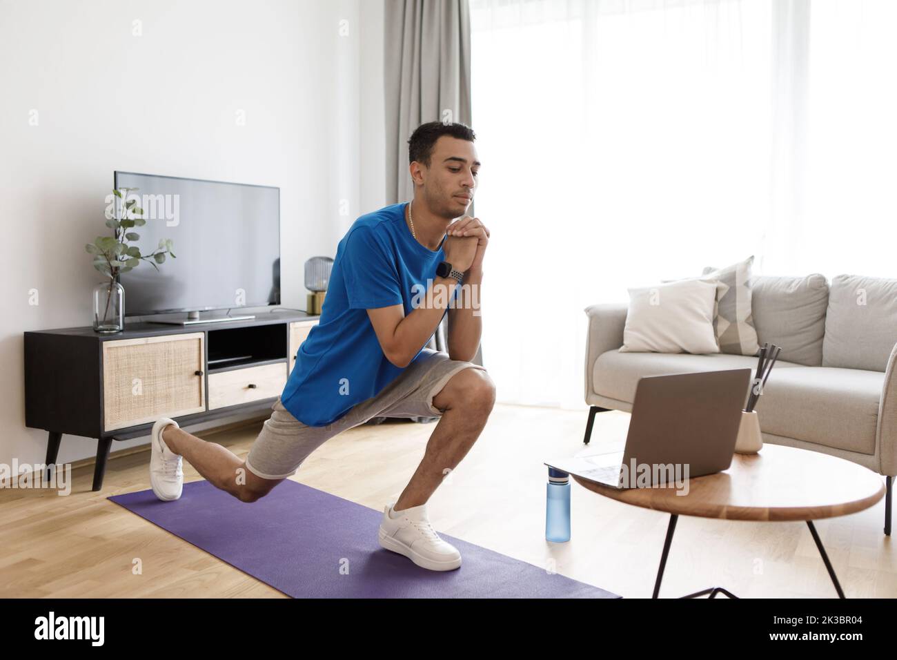 Jeune homme arabe regardant un tutoriel d'entraînement sur ordinateur portable et faisant des fentes de marche avant sur le tapis de fitness, s'exerçant à la maison Banque D'Images