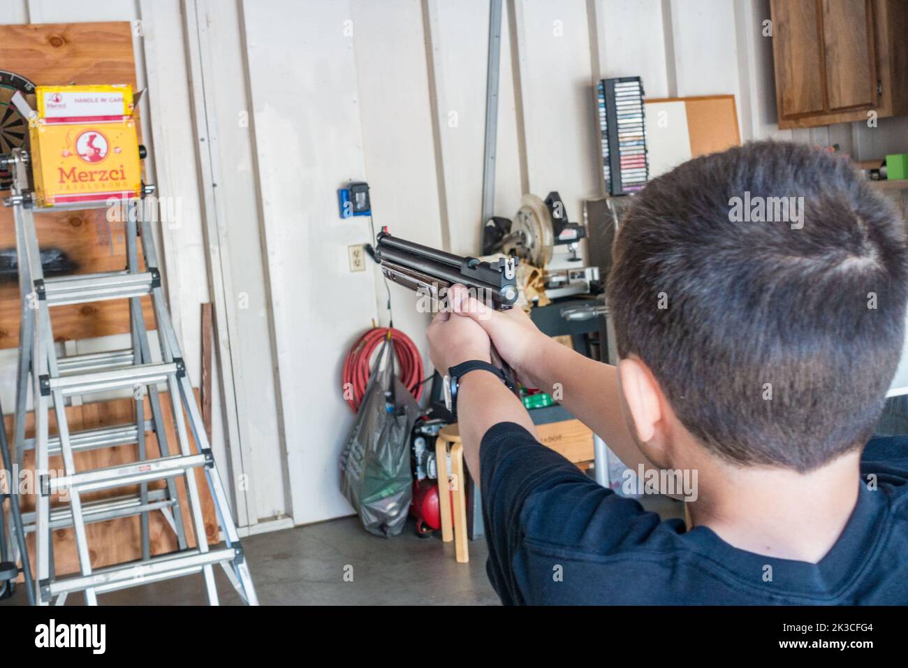 Vue sur l'épaule d'un garçon visant et tirant un pistolet à air dans un garage. Banque D'Images