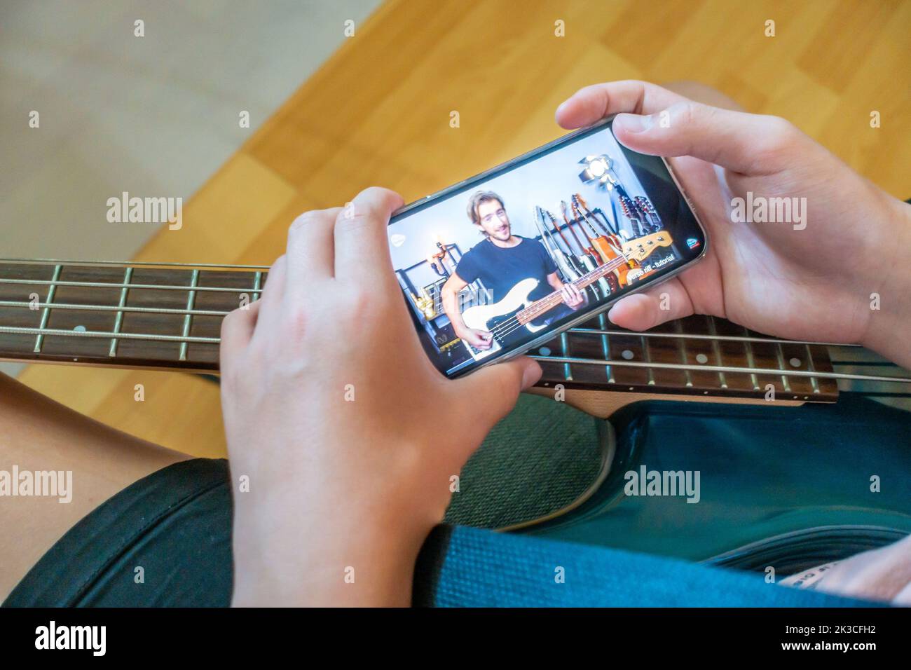 Un garçon auto-éduque en regardant une vidéo d'instruction sur un smartphone pour apprendre à jouer un morceau sur une guitare basse électrique Banque D'Images