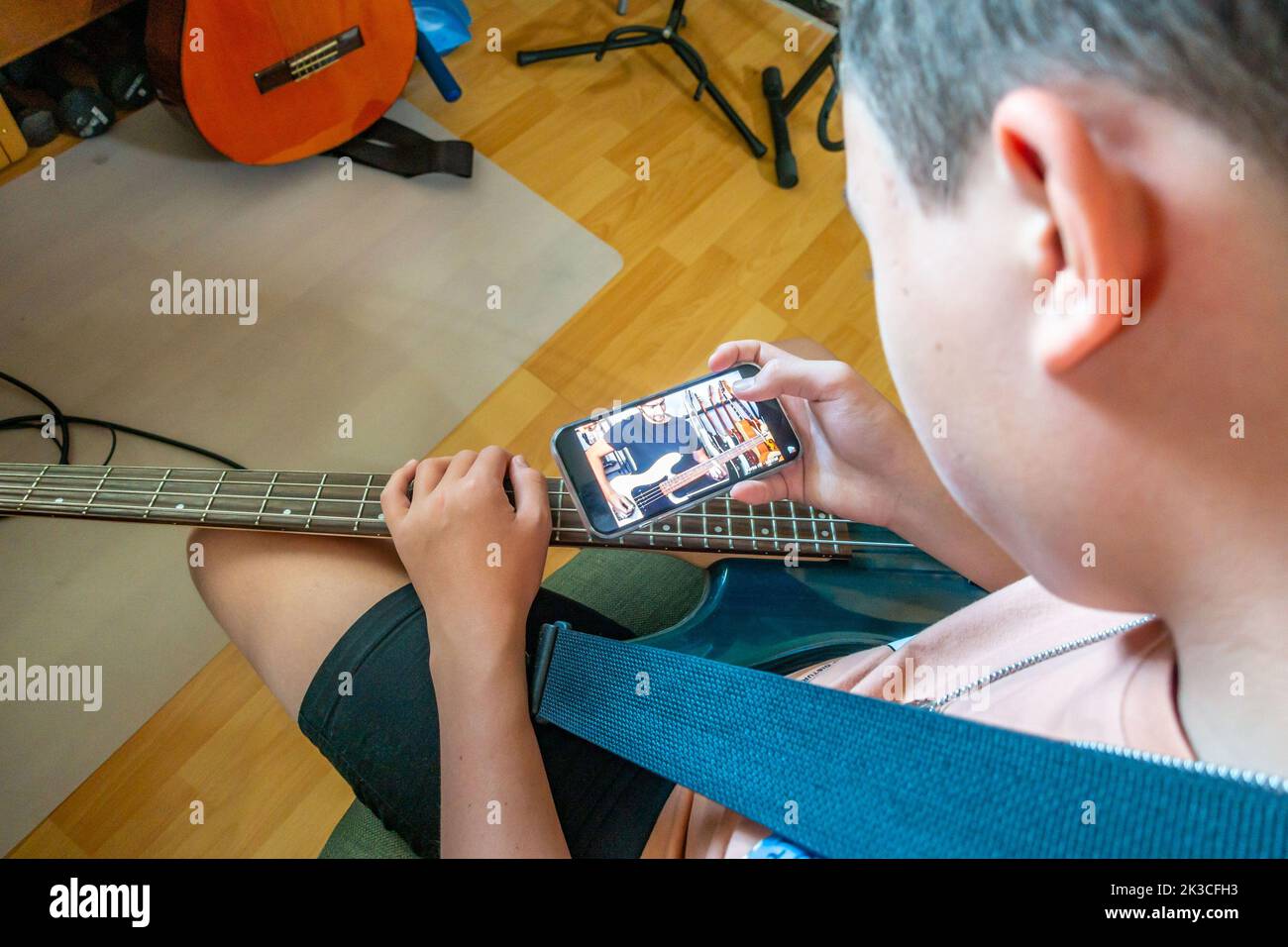 Un garçon auto-éduque en regardant une vidéo d'instruction sur un smartphone pour apprendre à jouer un morceau sur une guitare basse électrique Banque D'Images