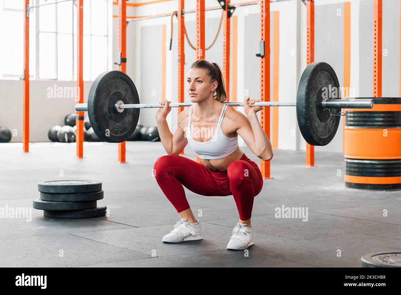Athlète féminine en plein corps en forme d'activewear regardant loin et faisant le dos de barbell squat pendant l'entraînement intense dans la salle de sport ensoleillée Banque D'Images