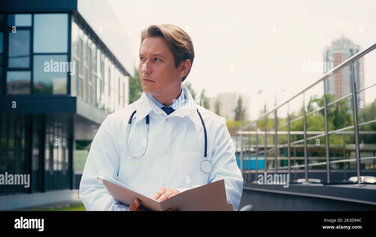 docteur en manteau blanc avec stéthoscope et dossier regardant loin à l'extérieur, image de stock Banque D'Images