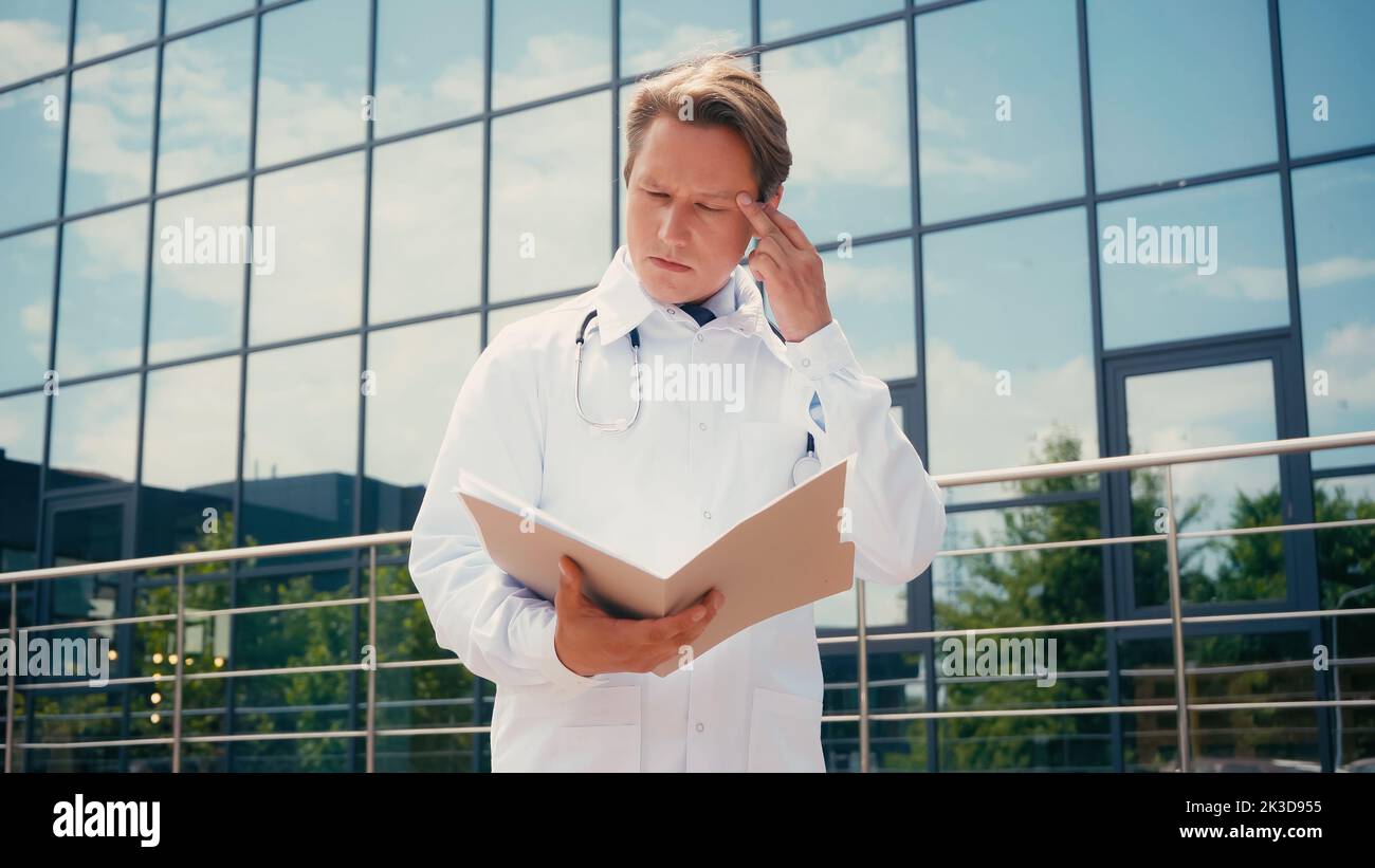 médecin attentif touchant la tête en regardant dans un dossier avec des documents à l'extérieur, image de stock Banque D'Images