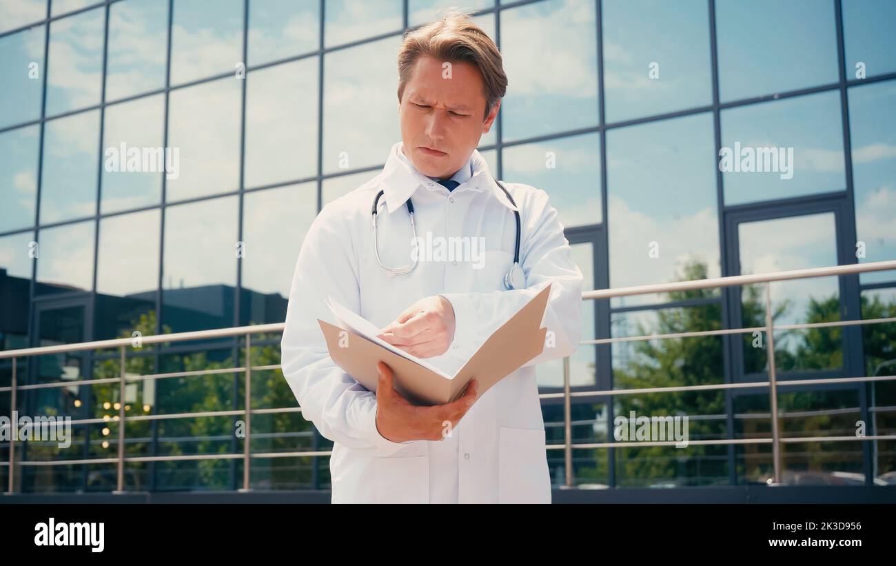 médecin attentif regardant dans un dossier avec des documents près de la clinique à l'extérieur, image de stock Banque D'Images
