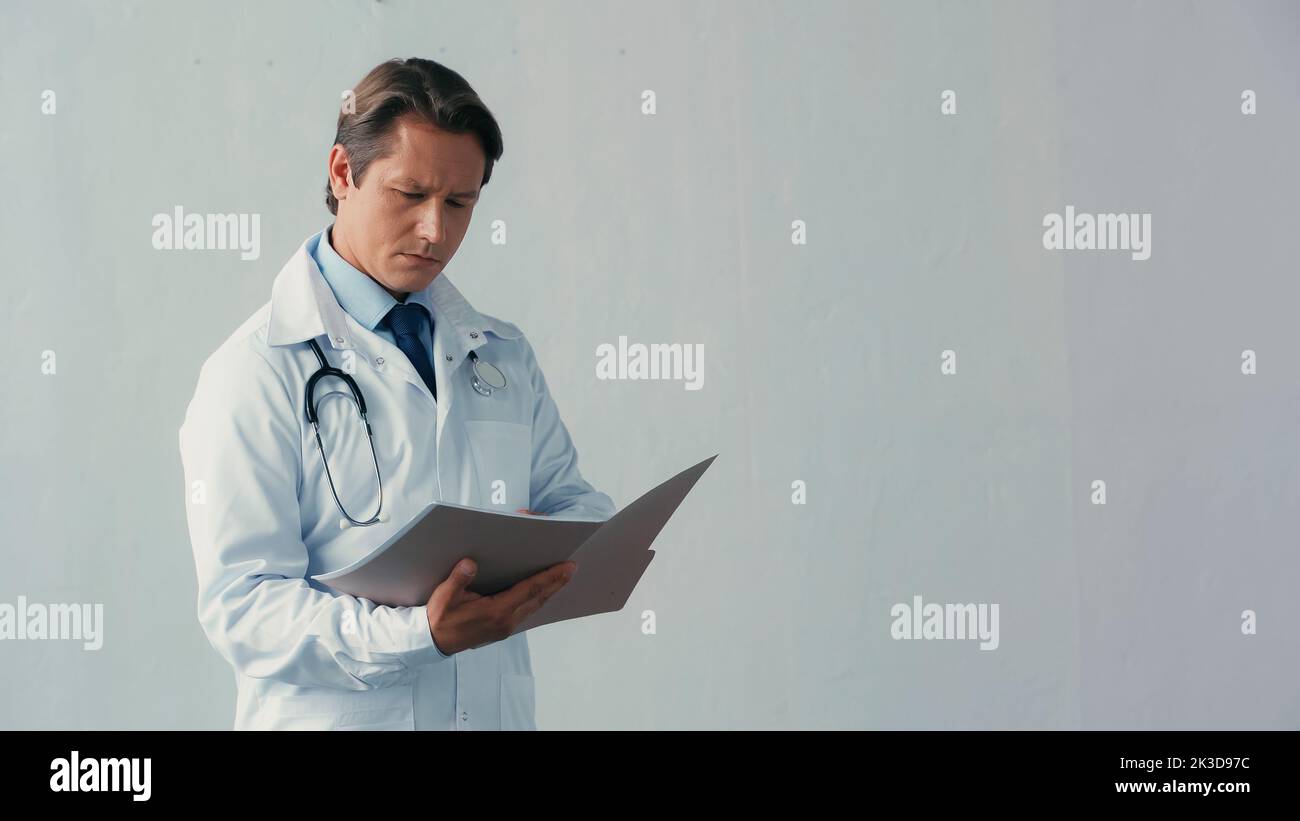 médecin attentif en manteau blanc regardant dans le dossier avec des documents sur gris, image de stock Banque D'Images
