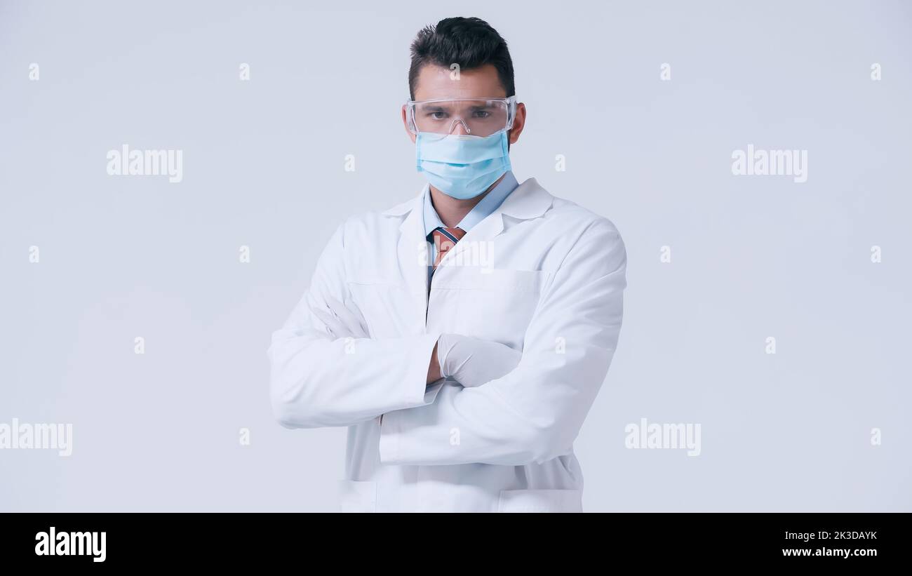 médecin en manteau blanc et masque médical avec des lunettes debout avec des bras croisés isolés sur image grise Banque D'Images