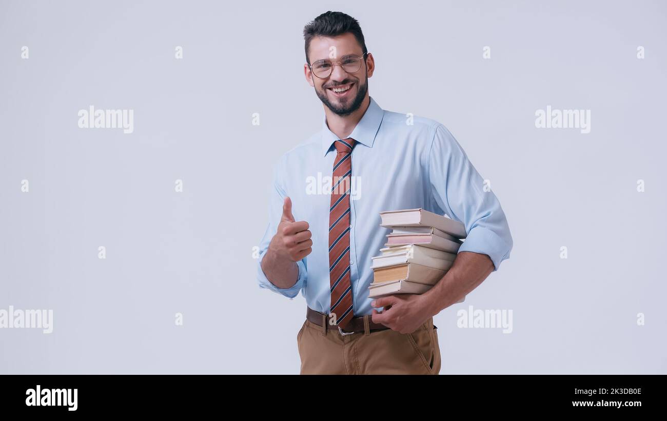 professeur heureux dans les lunettes tenant des manuels et montrant le pouce isolé sur gris, image de stock Banque D'Images