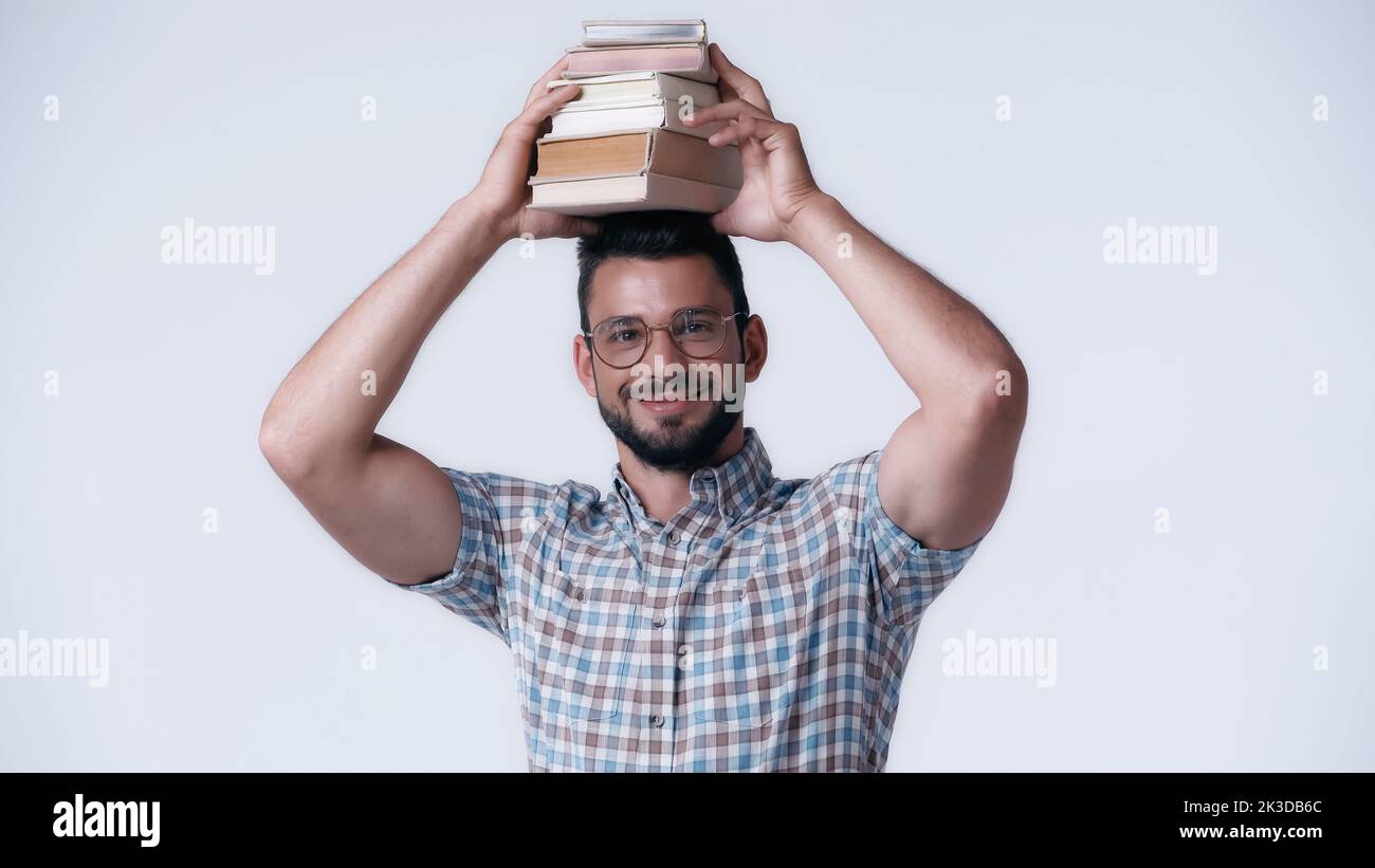 joyeux étudiant de nerd dans des lunettes tenant pile de livres au-dessus de la tête isolé sur gris, image de stock Banque D'Images