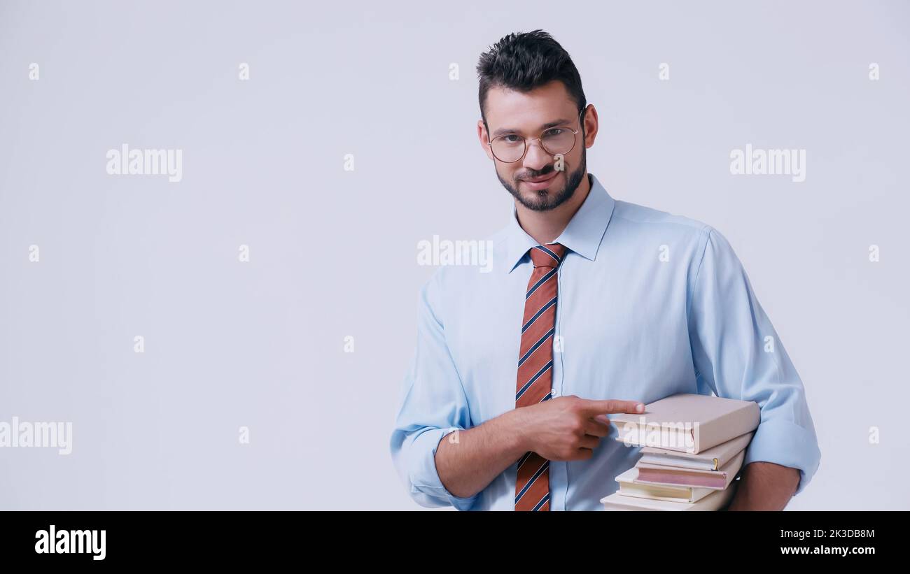 jeune professeur de lunettes pointant à la pile de livres isolés sur gris, image de stock Banque D'Images