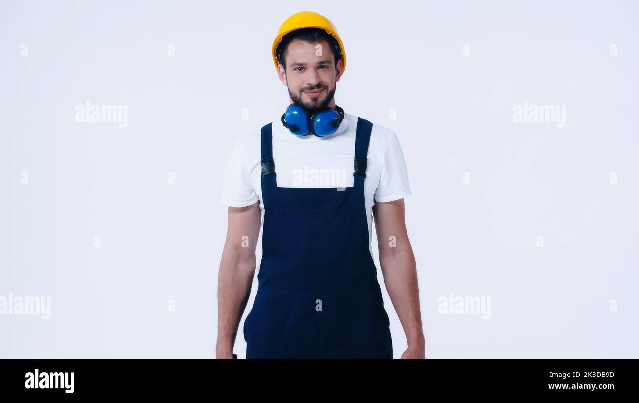 jeune bâtisseur en combinaison et casque de sécurité souriant à la caméra isolée sur blanc, image de stock Banque D'Images