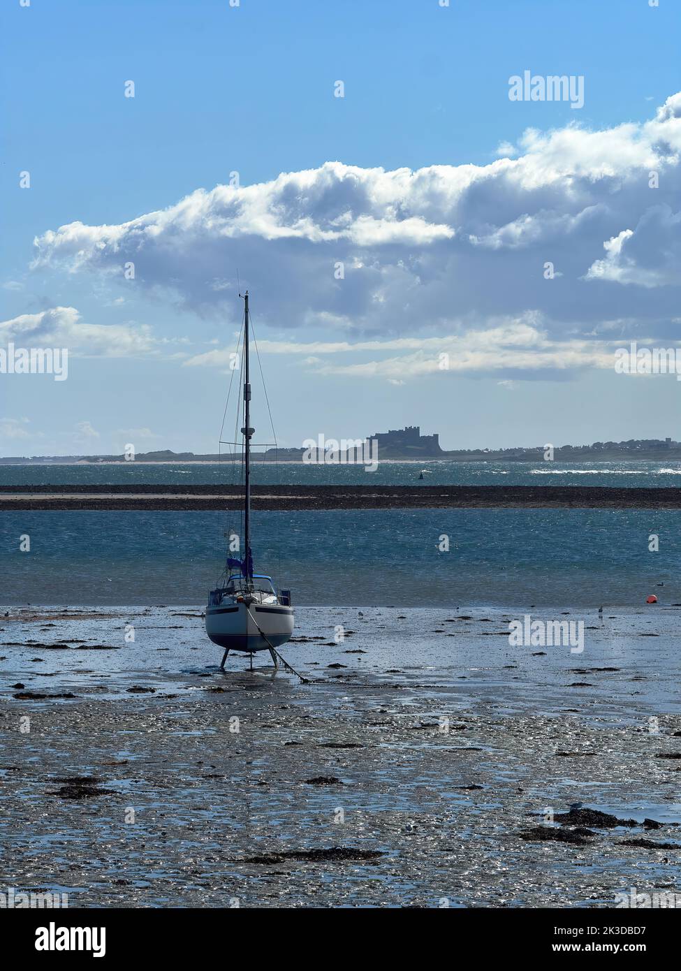 Un yacht perché sur sa quille dans la zone intertidale, avec la mer du Nord et le château de Bamburgh derrière et un paysage nuageux élaboré au-dessus. Banque D'Images