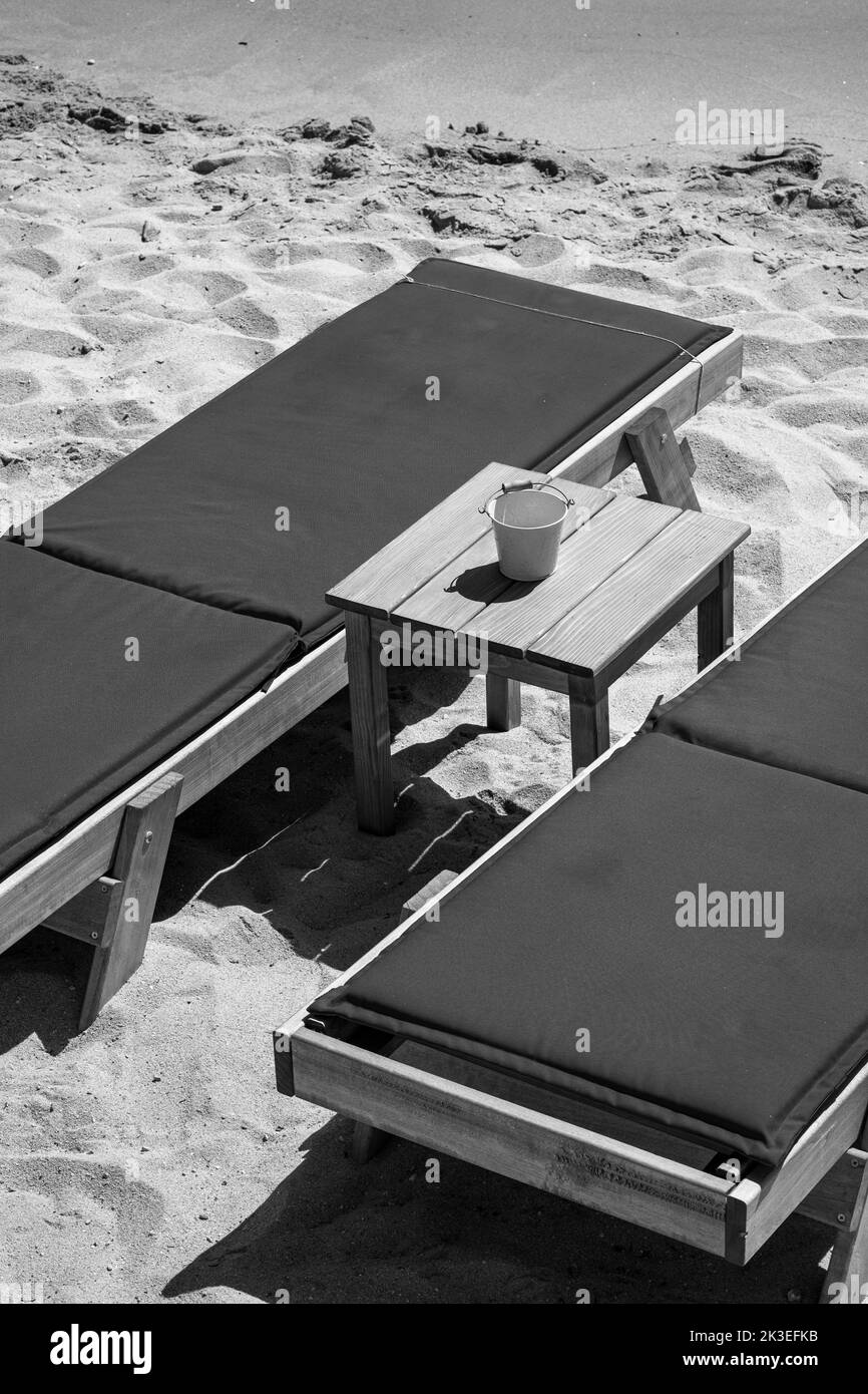 Vue rapprochée de deux chaises longues bleues et d'une petite table en face de la belle plage de sable doré de Mylopotas dans iOS Grèce en noir et blanc Banque D'Images