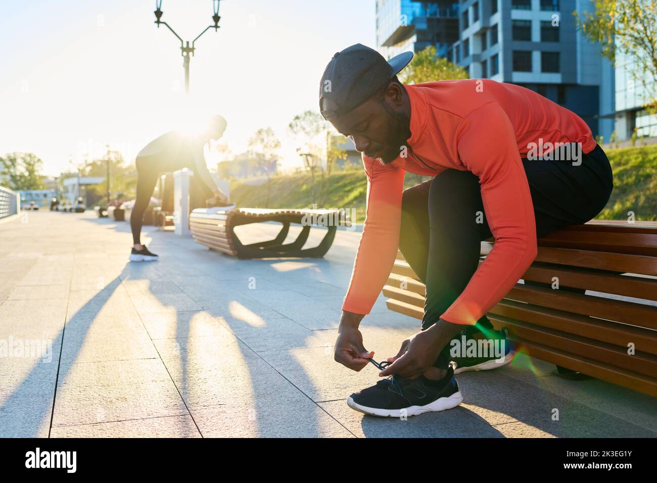 Jeune sportif afro-américain en veste de sport rouge et en leggins noirs qui noue le renfort de la sneaker tout en se pliant vers l'avant par-dessus la jambe Banque D'Images