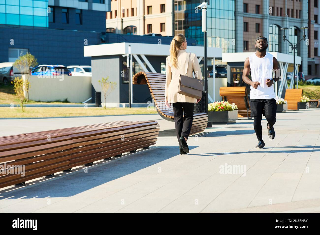 Un jeune sportif en forme d'activewear qui fait du jogging le long de la route dans le parc tandis qu'une femme d'affaires blonde en tenue de cérémonie le passant le matin Banque D'Images