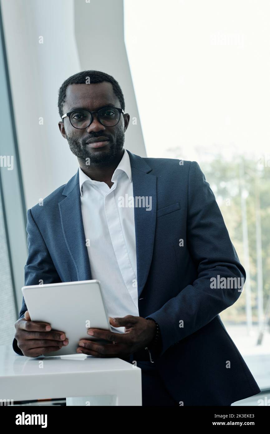 Jeune homme afro-américain confiant en costume et lunettes regardant la caméra tout en se tenant sur le lieu de travail et en utilisant une tablette Banque D'Images