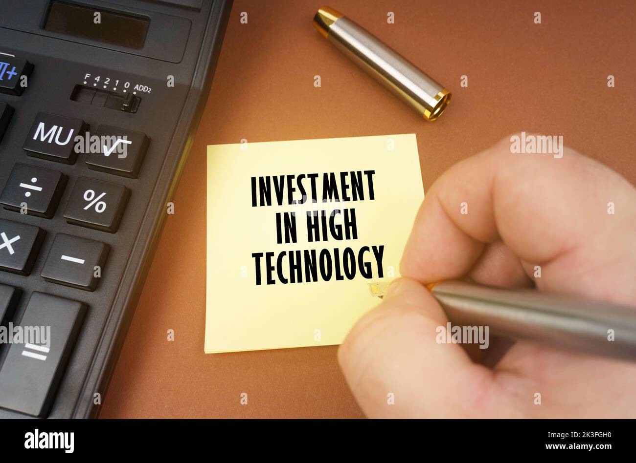 Concept d'entreprise. Une calculatrice repose sur une surface marron, une main avec un stylo fait une inscription sur un autocollant - investissement dans la haute technologie Banque D'Images