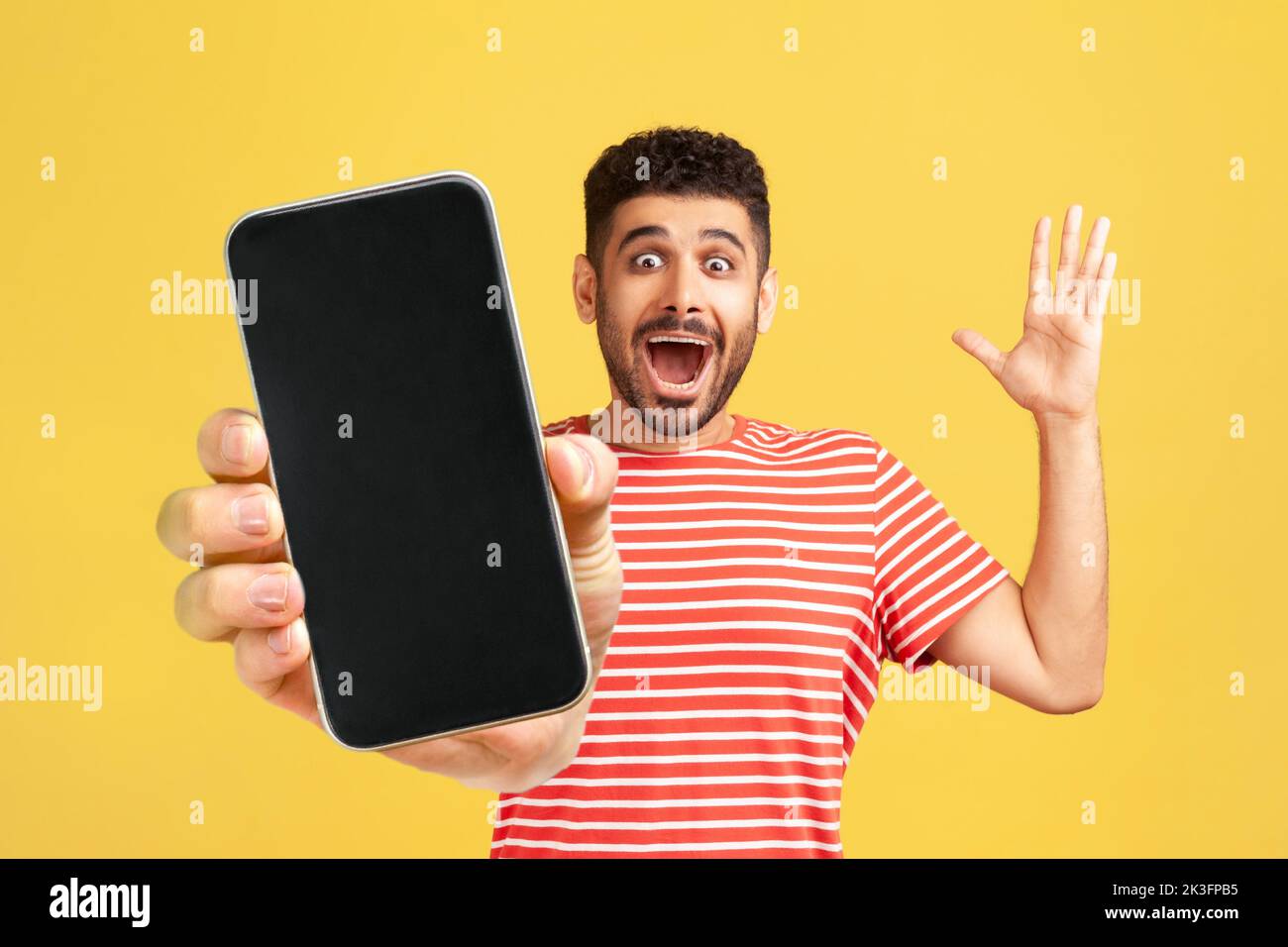 Un homme joyeux et émerveillé avec une barbe portant un T-shirt rouge montrant un téléphone portable avec écran vierge, exprimant le bonheur, le bras relevé et criant heureux. Studio d'intérieur isolé sur fond jaune. Banque D'Images Un homme joyeux et émerveillé avec une barbe portant un T-shirt rouge montrant un téléphone portable avec écran vierge, exprimant le bonheur, le bras relevé et criant heureux. Studio d'intérieur isolé sur fond jaune. Banque D'Images