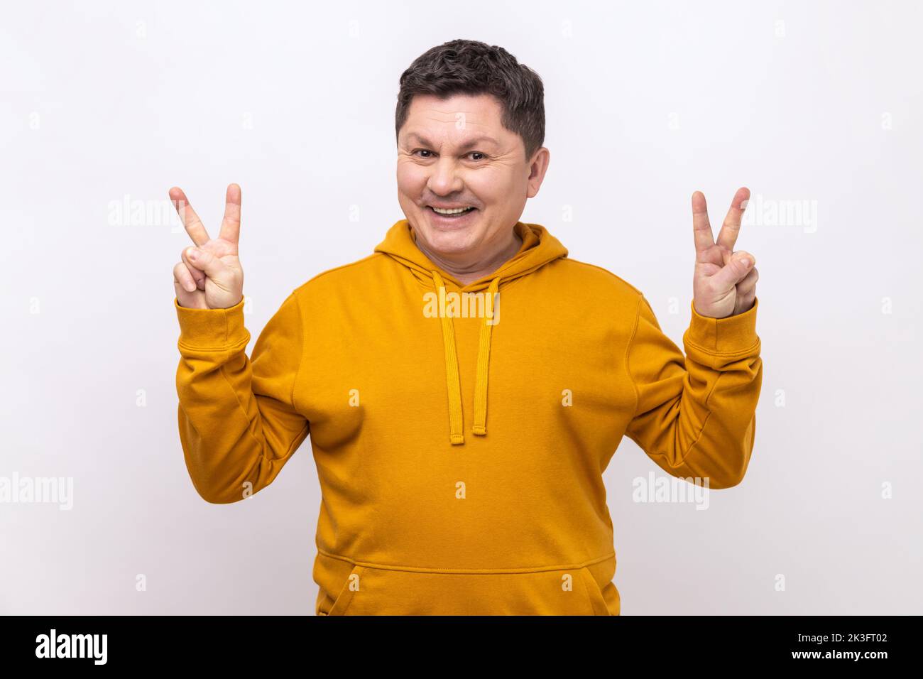 Joyeux positif homme d'âge moyen montrant le signe de v à l'appareil photo, en se vetant et souriant, homme souhaitant bonne chance, la paix, portant le style urbain de sweat à capuche. Studio d'intérieur isolé sur fond blanc. Banque D'Images