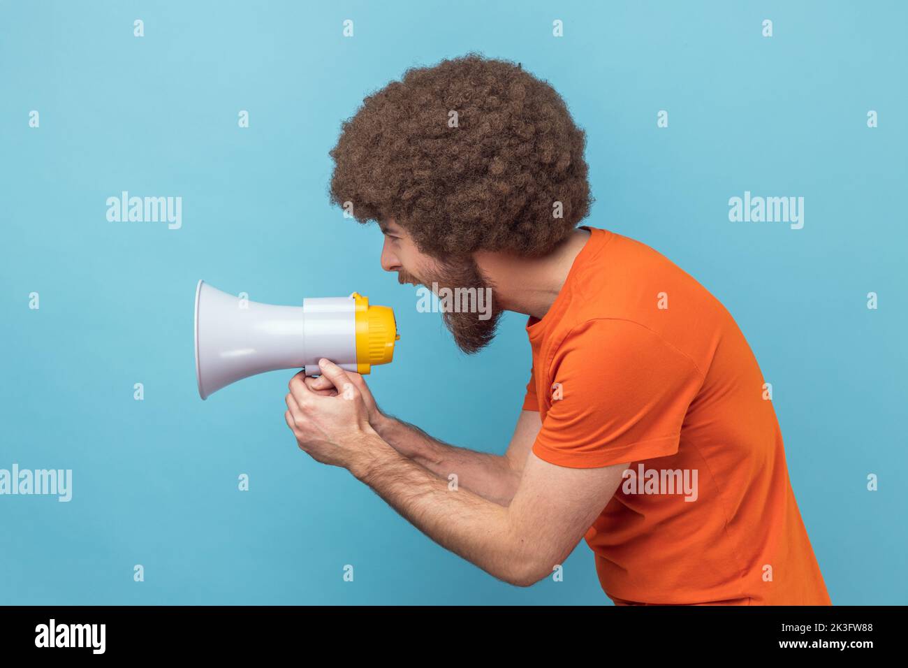 Vue latérale d'un homme avec une coiffure afro portant un T-shirt orange criant bruyamment au mégaphone, faisant annoncer, protestant, veut être entendu. Studio d'intérieur isolé sur fond bleu. Banque D'Images