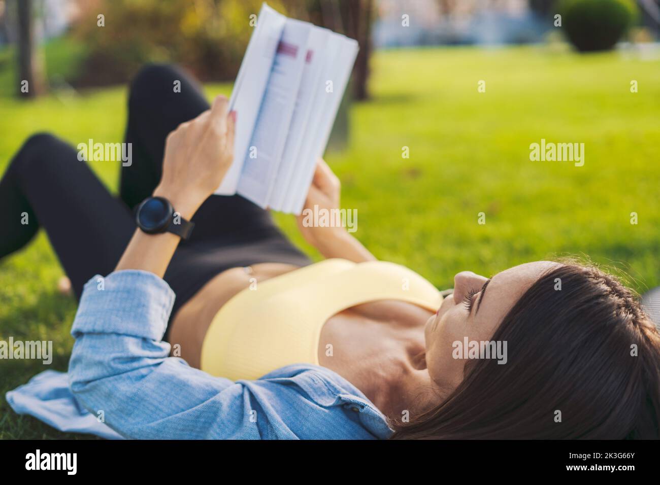 Belle jeune femme sportive lisant un livre dans le parc allongé sur le tapis de yoga, matin détente. Concept : donner le temps pour vous-même de lire des livres Banque D'Images
