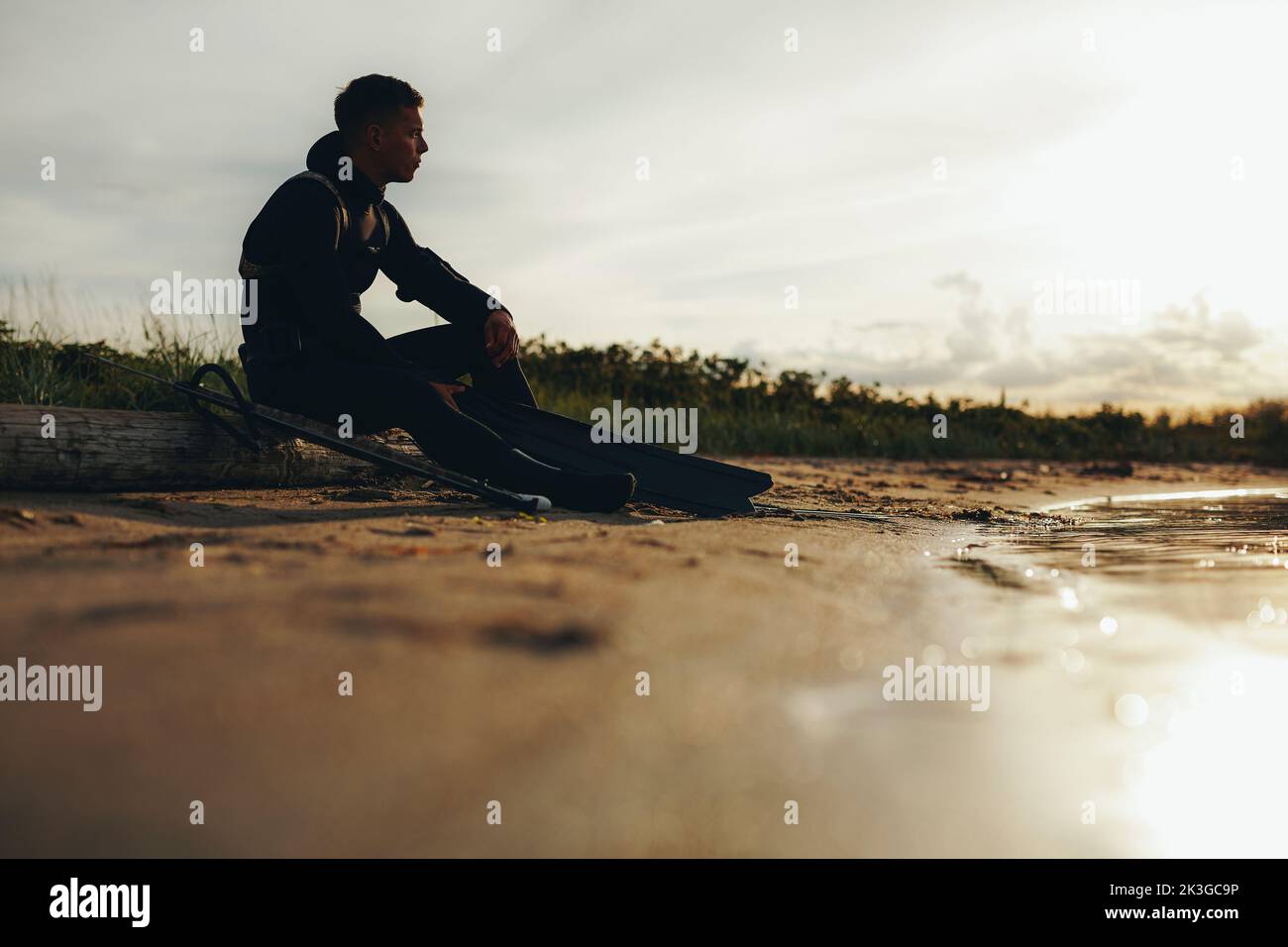 Jeune plongeur masculin assis sur la plage avec un fer de lance et du matériel de pêche. Un jeune homme aventureux qui regarde le coucher du soleil avec attention. Banque D'Images