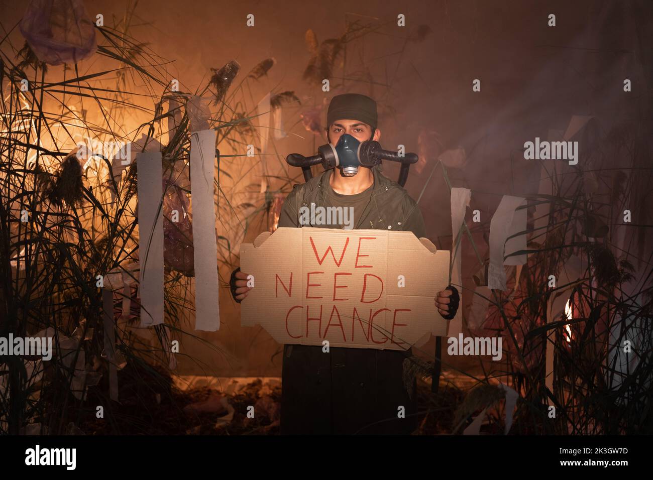 Un homme dans un masque à gaz tenant une affiche avec des mots nous avons besoin de changement. Brûler des déchets en plastique sur le fond. Problèmes environnementaux: Déchets en plastique Banque D'Images
