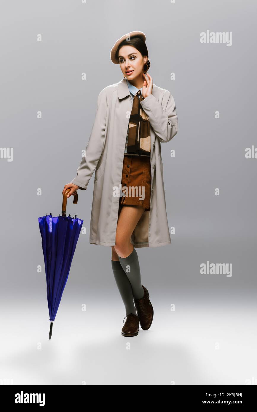 pleine longueur de jeune femme élégante en béret et trench manteau posant avec parapluie bleu sur gris, image de stock Banque D'Images