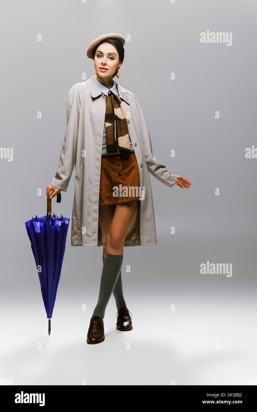 pleine longueur de brune jeune femme en béret et trench manteau posant avec parapluie bleu sur gris, image de stock Banque D'Images