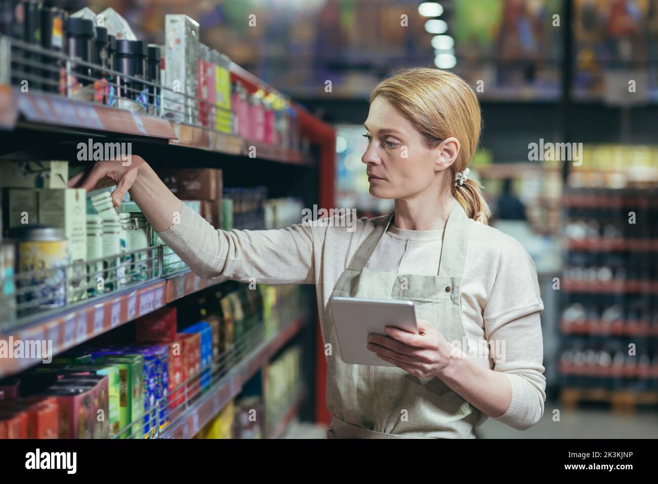 Une belle jeune femme employée d'un supermarché, stocker dans un uniforme de travail affiche les marchandises sur l'étagère, compte, écrit dans un carnet. Banque D'Images