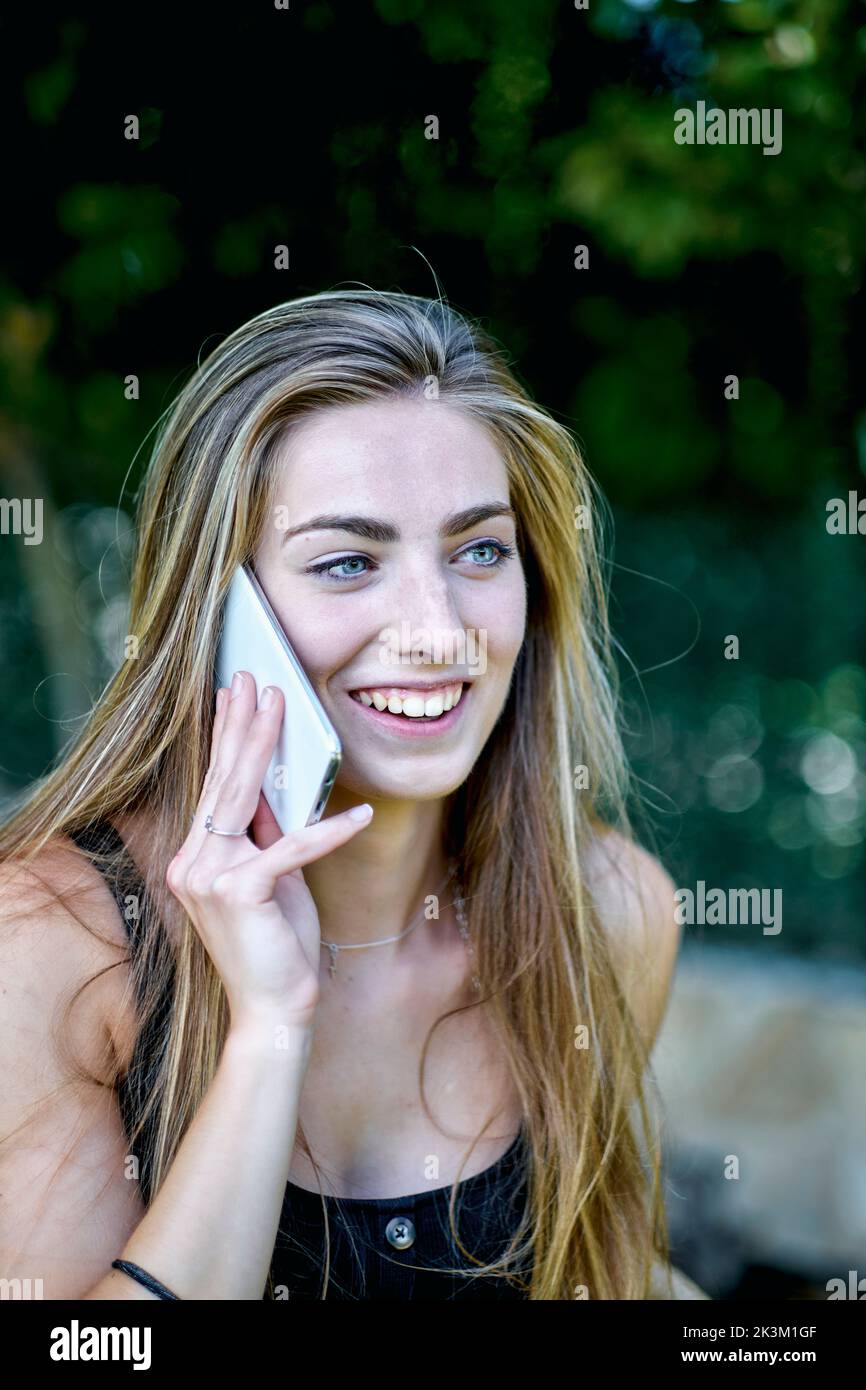 Portrait de beauté naturelle d'une jeune femme dans ses années 20 avec de longs cheveux et des yeux bleus à l'extérieur dans un jardin parlant sur son téléphone mobile. Concept de style de vie. Banque D'Images