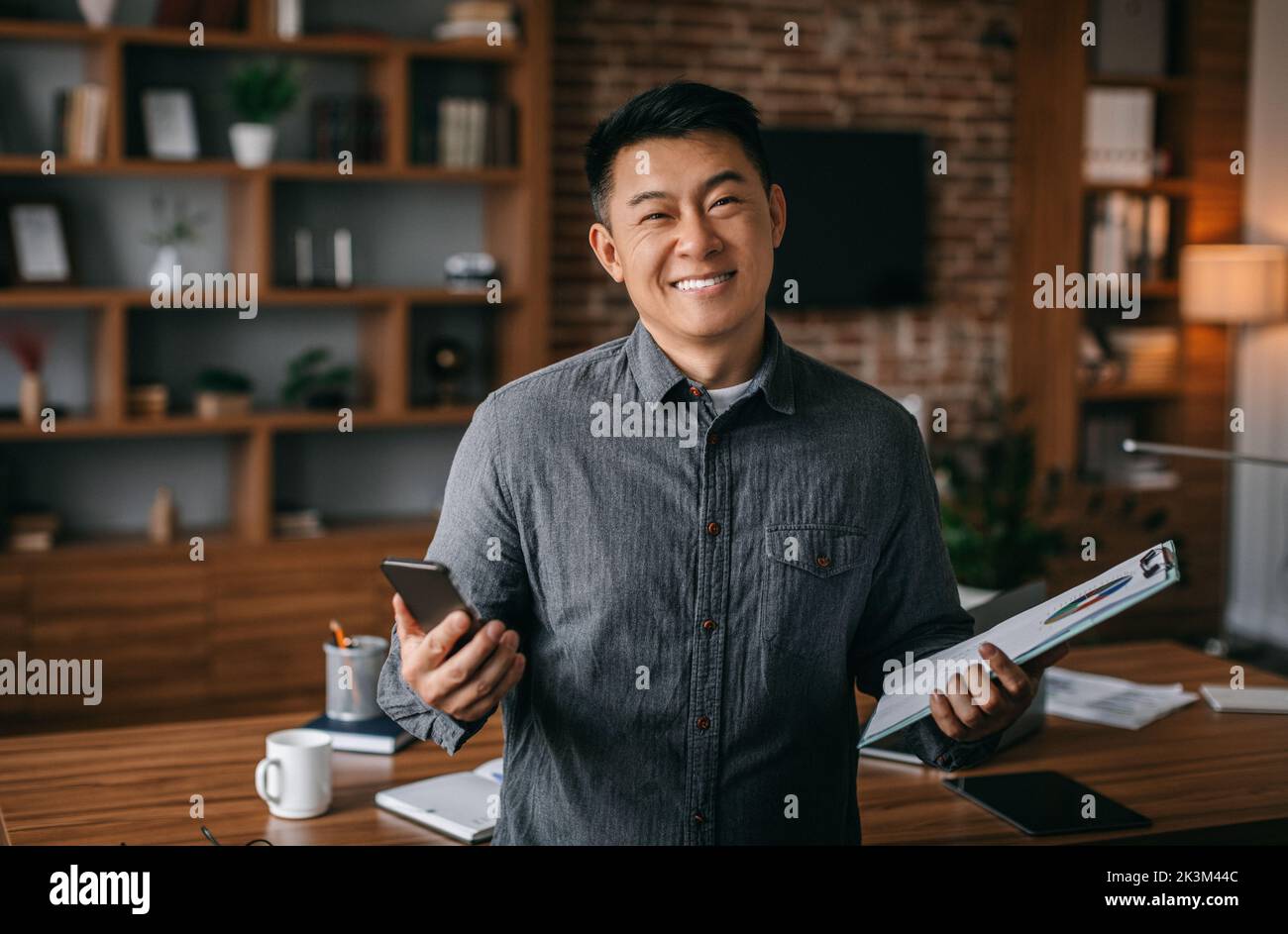 Homme d'affaires travaille à la maison. Joyeux homme chinois d'âge moyen réussi avec tablette et téléphone à l'intérieur du bureau Banque D'Images