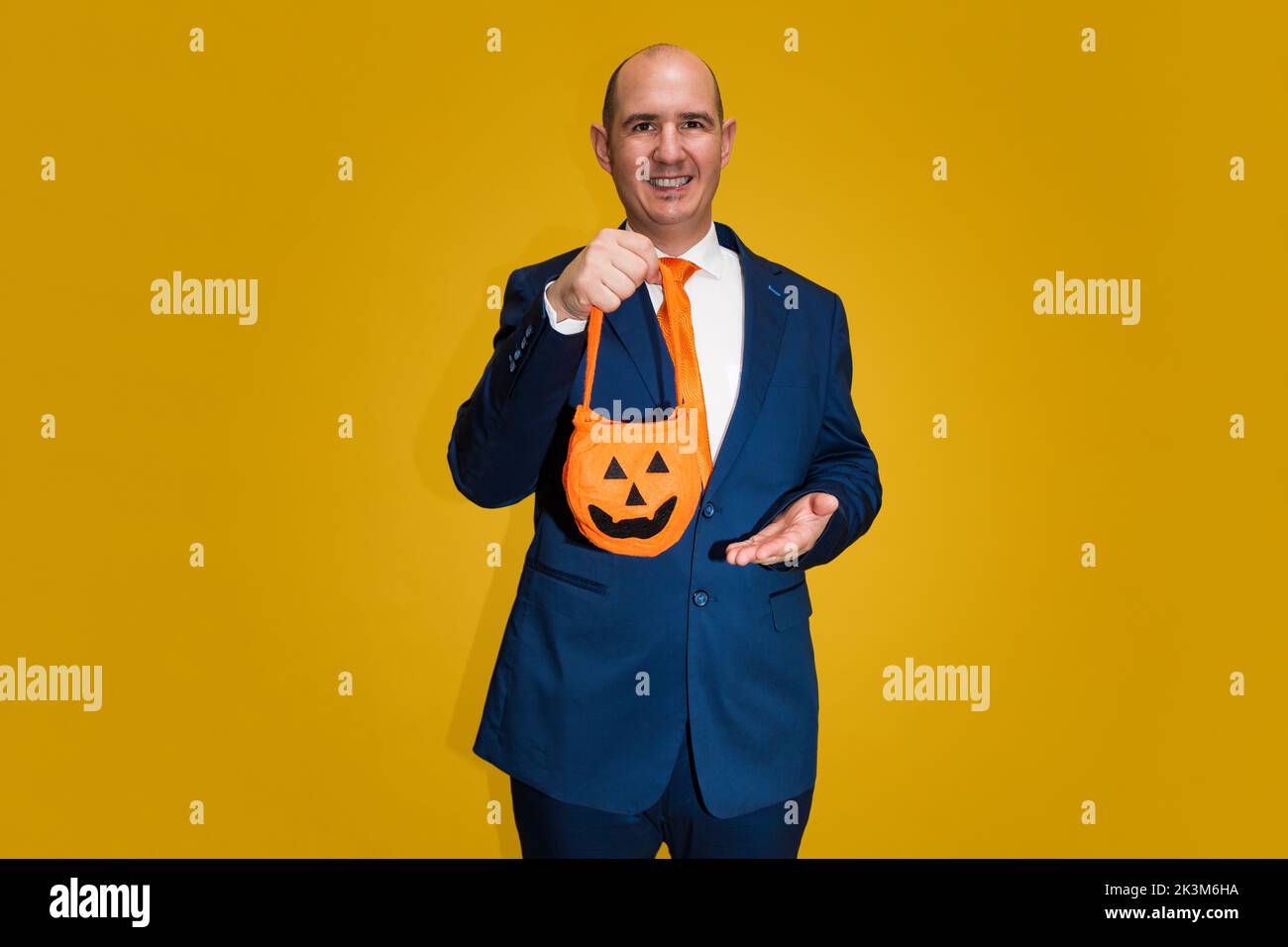 Un homme cacaucasien à tête blanche vêtu d'un costume bleu, d'une chemise blanche et d'une cravate orange tient un sac en tissu en forme de citrouille d'Halloween. Le backgro Banque D'Images