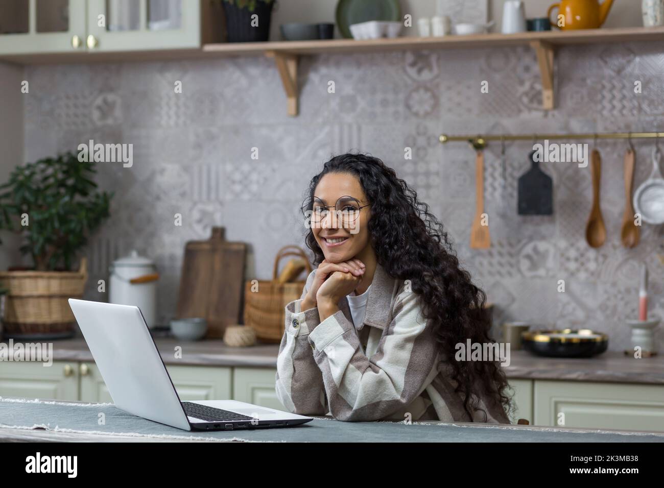 Portrait d'une jeune belle femme hispanique à la maison, femme indépendante travaillant à distance à l'aide d'un ordinateur portable, regardant l'appareil photo et souriant dans des lunettes et des cheveux bouclés dans la cuisine Banque D'Images