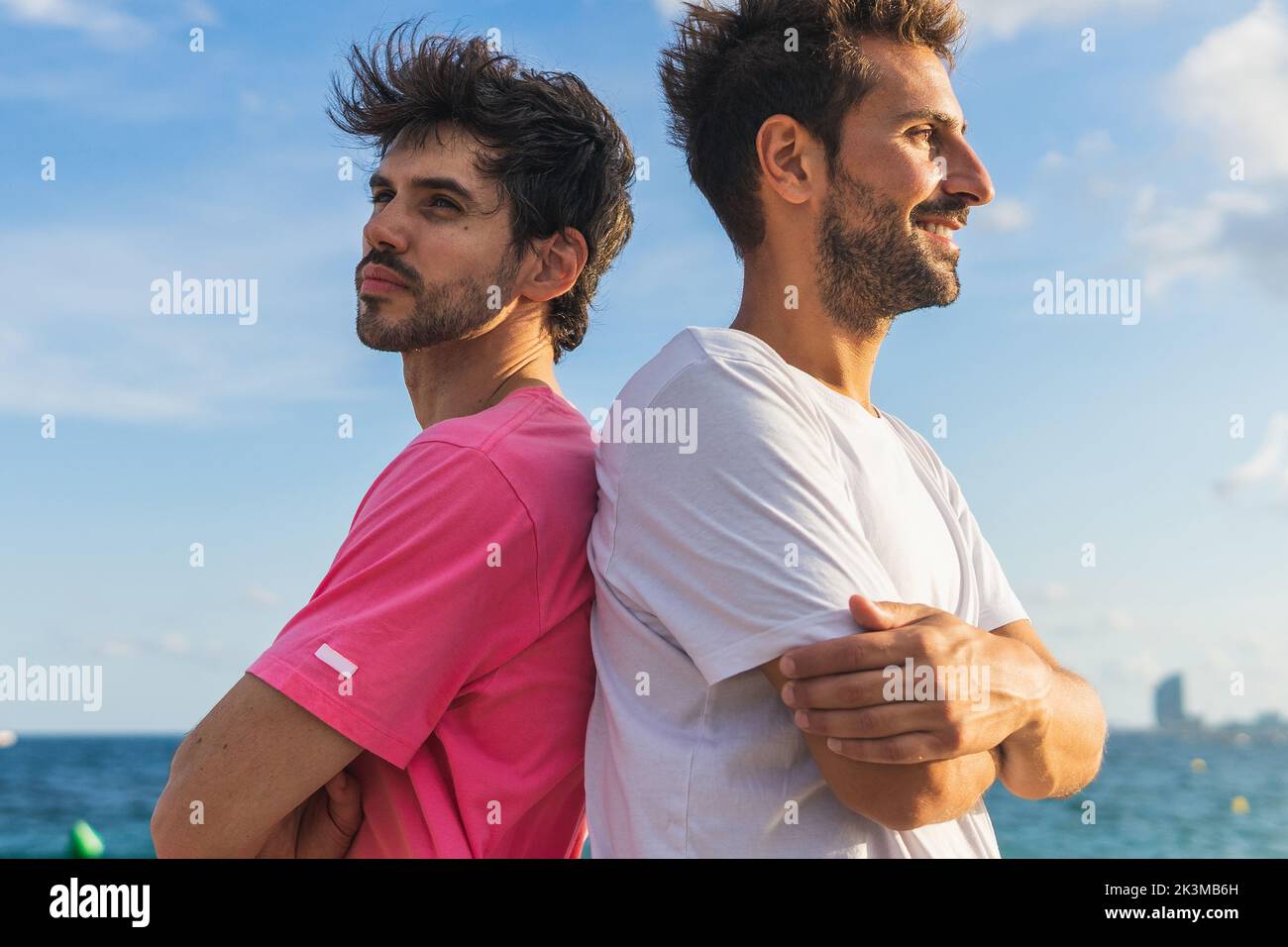 Des athlètes hispaniques en t-shirts croisant les bras et regardant loin tout en se tenant dos à dos contre la mer et le ciel bleu pendant la pause dans fitne Banque D'Images Des athlètes hispaniques en t-shirts croisant les bras et regardant loin tout en se tenant dos à dos contre la mer et le ciel bleu pendant la pause dans fitne Banque D'Images