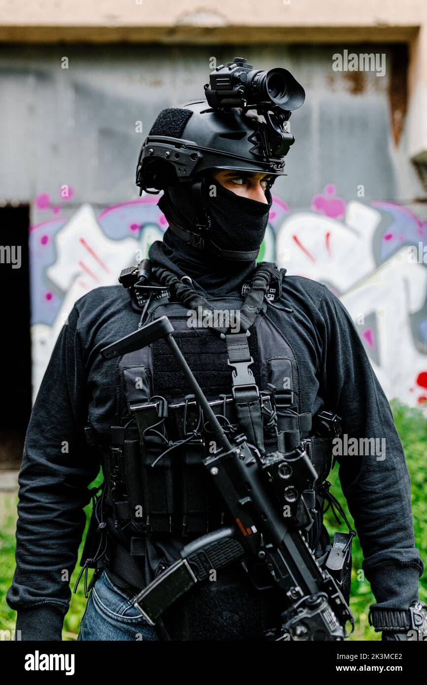 Jeune guerrier masculin méconnaissable, sérieux et méconnaissable, porter une balaclava moderne et un casque avec une carabine professionnelle qui s'éloigne tout en se tenant près de l'ancien Banque D'Images