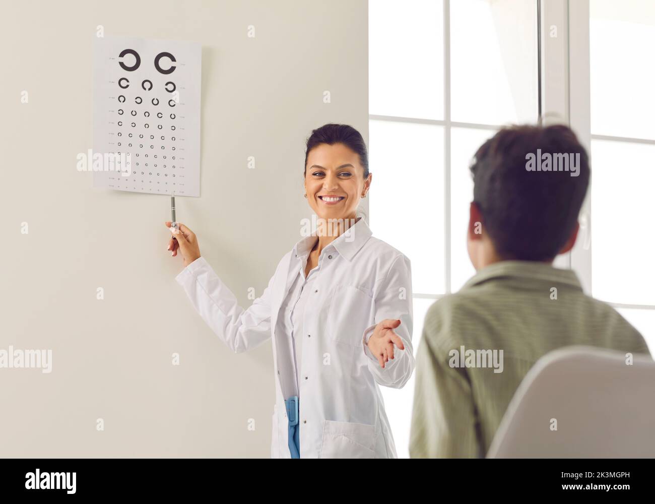 Ophtalmologiste souriant testant la vue de l'enfant et pointant vers le diagramme des yeux sur le mur Banque D'Images