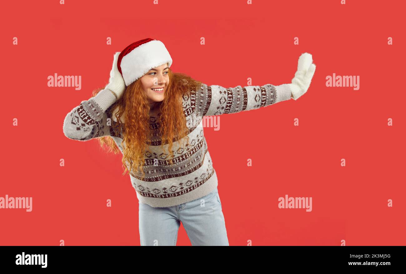 Femme heureuse en chandail, chapeau de Noël et moufles dansant isolée sur fond rouge Banque D'Images