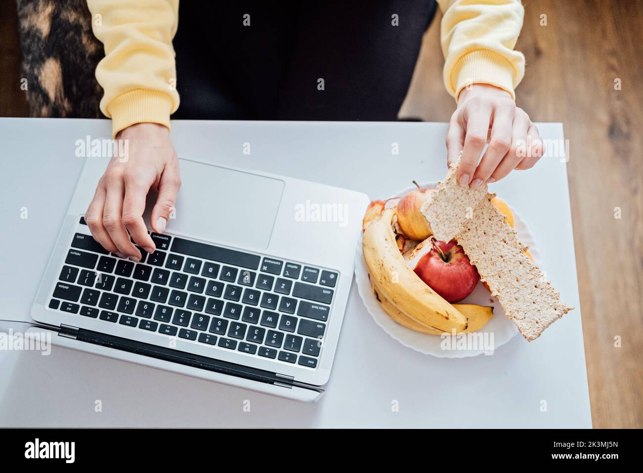Jeune femme indépendante qui mange des aliments sains lorsqu'elle travaille à la maison. Femme mangeant des collations et des fruits à grain sain tout en travaillant avec un ordinateur portable à la maison Banque D'Images