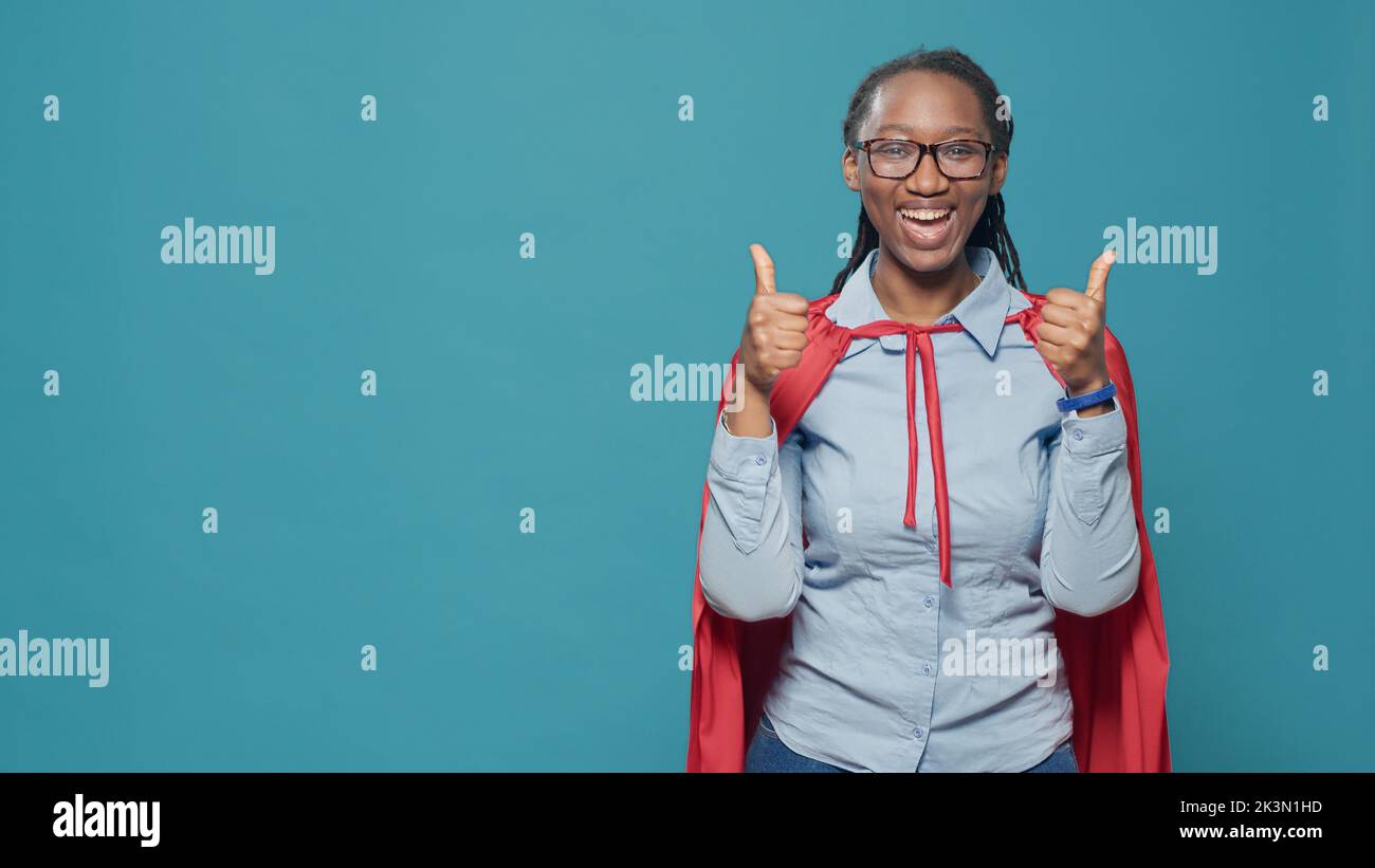 Femme super-héros avec cape faisant le pouce vers le haut geste sur l'appareil photo, comme la publicité et l'approbation signe avec le cloak rouge et héros de personnage de bande dessinée costume. Femme donnant bon symbole. Banque D'Images