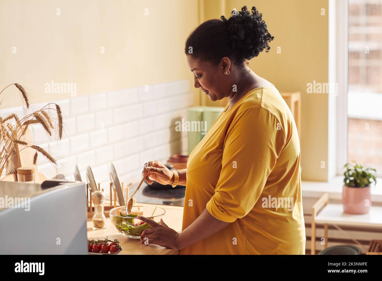Vue latérale portrait d'une femme noire en surpoids cuisant un repas sain dans la cuisine éclairée par la lumière du soleil, espace de copie Banque D'Images