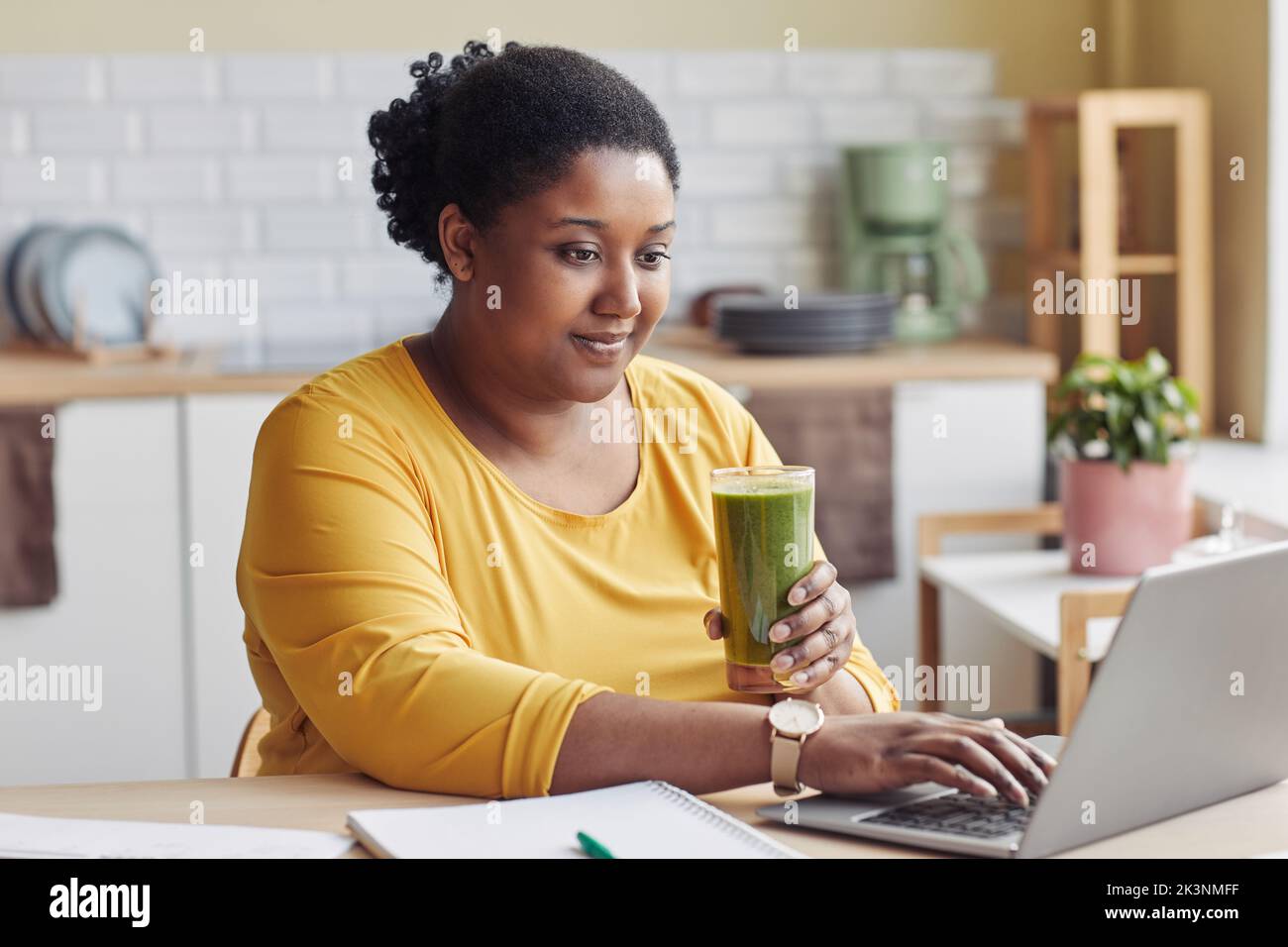 Portrait d'une femme noire en surpoids qui boit du smoothie et utilise un ordinateur portable à la maison Banque D'Images