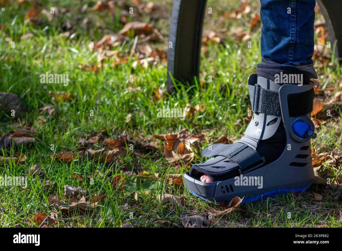 Un garçon avec un pied cassé et une chaussure orthopédique ou un marcheur après une fracture osseuse repose dans un parc public sur un banc en herbe verte pour recréer et réhabiliter Banque D'Images