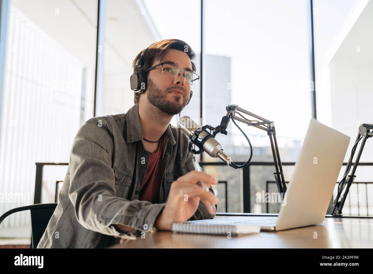 Jeune homme hôte dans des écouteurs et des lunettes assis à table, balayant le podcast audio en utilisant le microphone et l'ordinateur portable en studio, tenant un stylo, regardant vers. Médias sociaux, podcasting, concept de blogging Banque D'Images