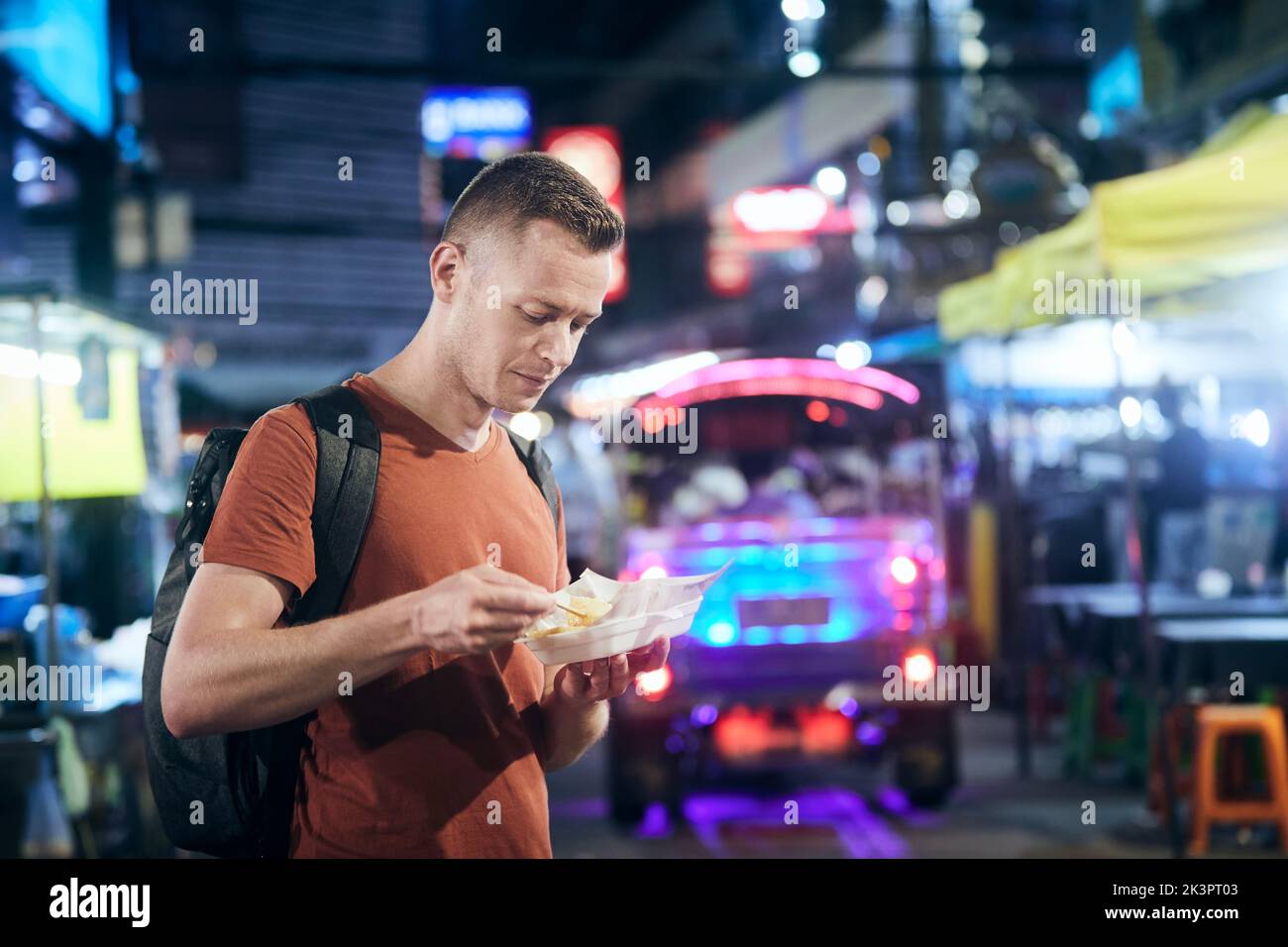 Tourisme de nuit à Bangkok. Homme avec sac à dos manger de la nourriture locale au marché de rue. Banque D'Images
